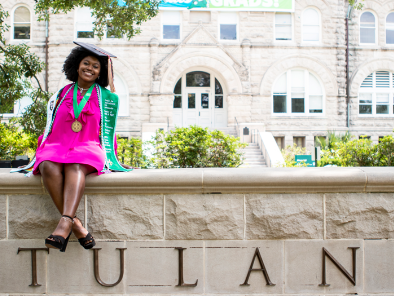 A Black woman in a pink dress and a graduation cap poses on a Tulane sign