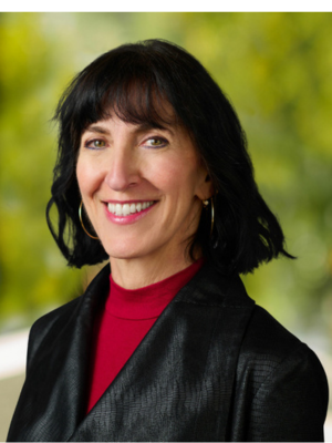 White woman in red shirt and black blazer smiles in front of tree